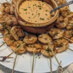 A plate of grilled shrimp on skewers encircles a central brown bowl filled with creamy dip, garnished with herbs, on a speckled countertop.