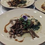 A gourmet dish featuring sautéed mushrooms, a meat portion, and greens garnished with a purple flower. Elegant presentation on a creamy sauce, set in a dining setting.