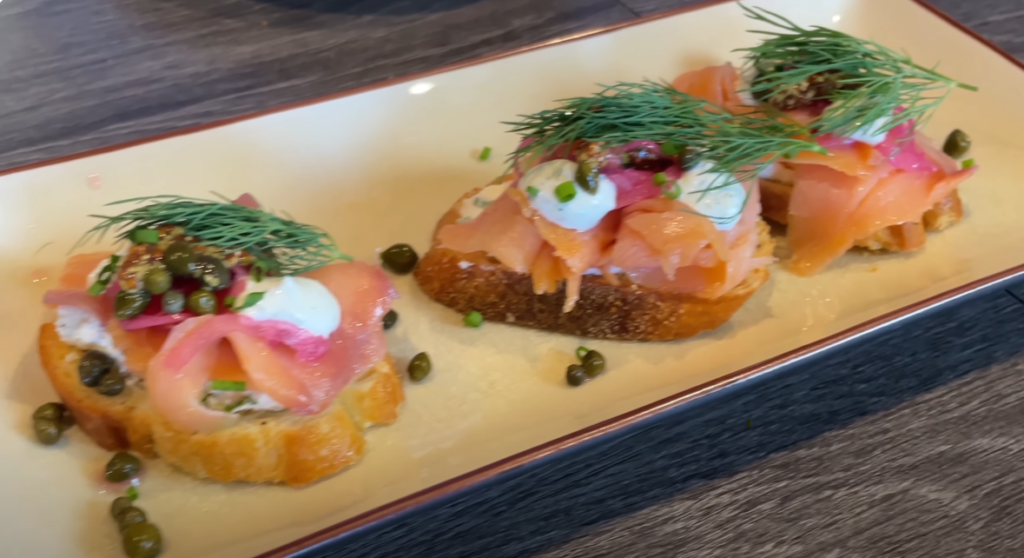Three pieces of bruschetta topped with smoked salmon, capers, dill, and cream cheese on a white plate. The dish looks fresh and appetizing.