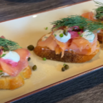 Three pieces of bruschetta topped with smoked salmon, capers, dill, and cream cheese on a white plate. The dish looks fresh and appetizing.