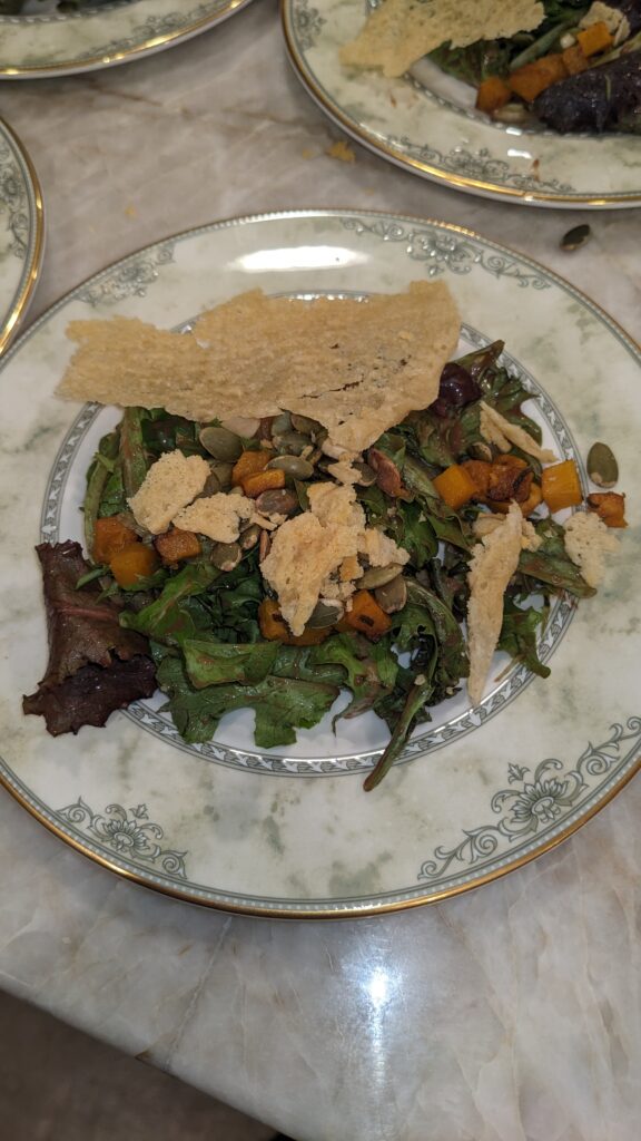A salad with mixed greens, roasted butternut squash, pumpkin seeds, and crispy crackers on a decorative white plate with a floral border.