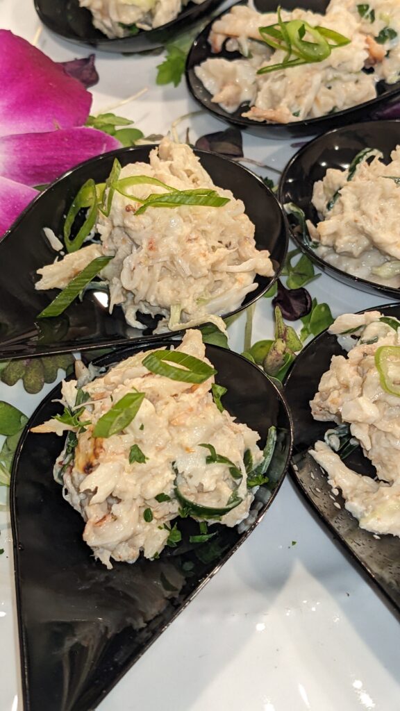Elegant crab salad served on black spoons, garnished with fresh herbs. A vibrant pink flower adds a touch of color, creating an appetizing presentation.