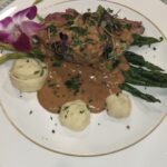 Gourmet dish on a white plate: steak with gravy, asparagus, mashed potato swirls, garnished with microgreens and an orchid, elegant presentation.