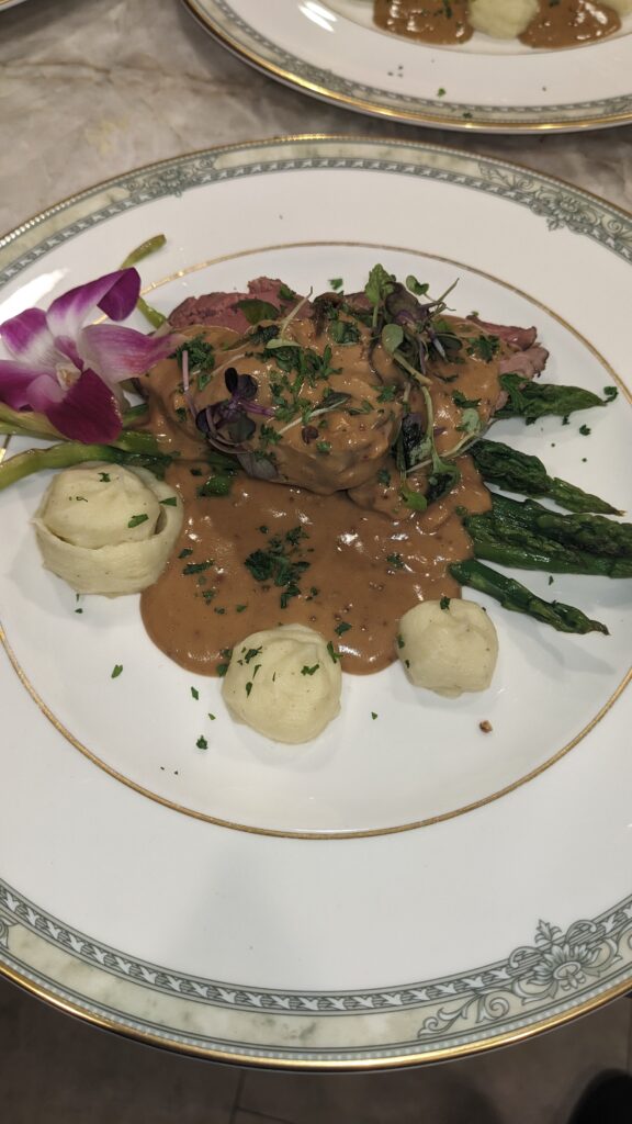 Gourmet dish on a white plate: steak with gravy, asparagus, mashed potato swirls, garnished with microgreens and an orchid, elegant presentation.