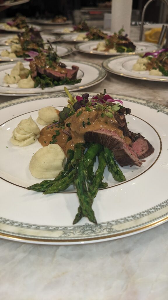 Plated gourmet dish featuring sliced steak with creamy sauce, asparagus, and mashed potatoes, garnished with edible flowers, exuding an elegant dining atmosphere.