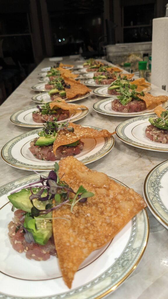 Elegant plates of tuna tartare topped with avocado and microgreens, paired with crispy wonton chips, lined up on a marble counter. Cozy ambiance.