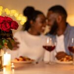 A romantic dinner scene shows a blurred couple smiling at each other. In focus are red roses, two glasses of wine, and lit candles, creating a warm ambiance.