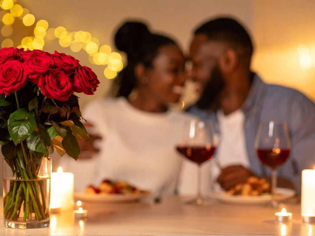 A romantic dinner scene shows a blurred couple smiling at each other. In focus are red roses, two glasses of wine, and lit candles, creating a warm ambiance.
