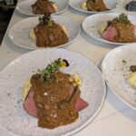 Six plates of gourmet beef, each served with mashed potatoes, rich brown sauce, and microgreens, arranged neatly on a white table.