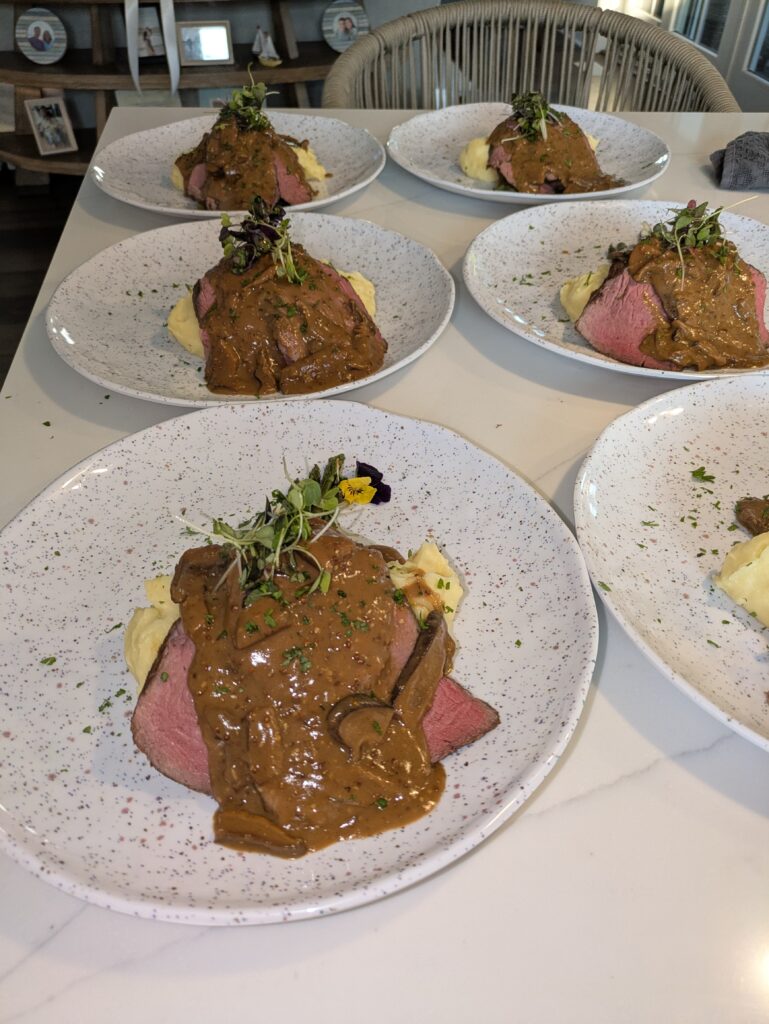 Six plates of gourmet beef, each served with mashed potatoes, rich brown sauce, and microgreens, arranged neatly on a white table.