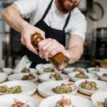 A chef seasons dishes with a pepper grinder in a bustling kitchen. Plates of elegant food, garnished with herbs and prosciutto, ready for serving.