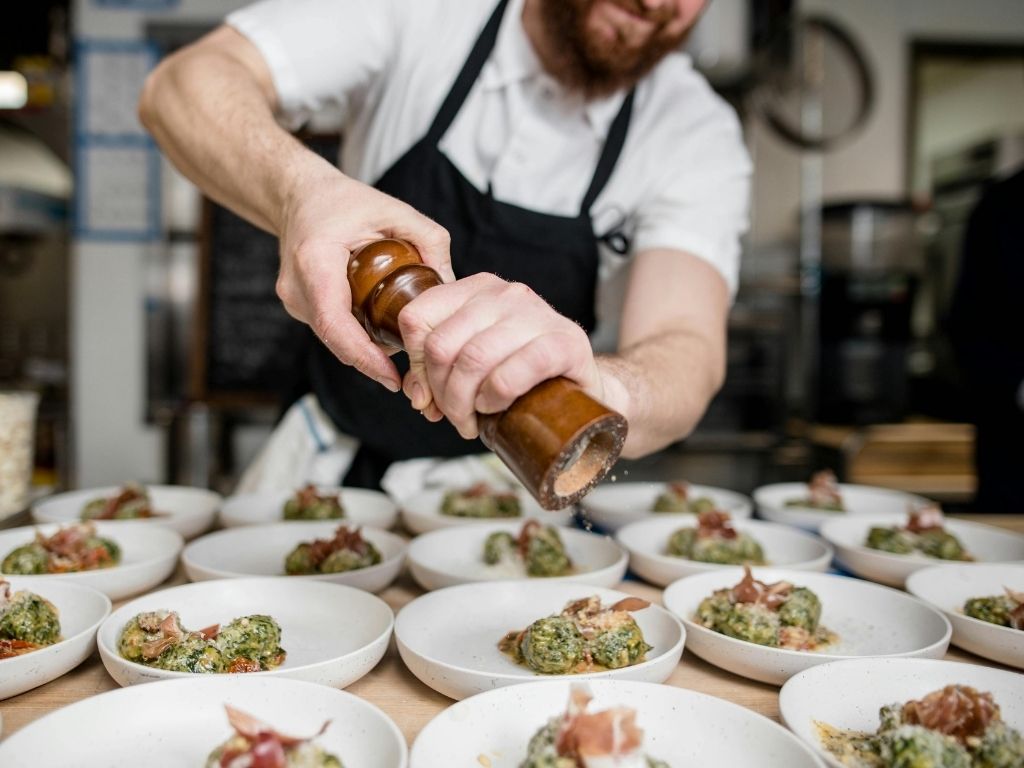 A chef seasons dishes with a pepper grinder in a bustling kitchen. Plates of elegant food, garnished with herbs and prosciutto, ready for serving.