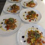 Eight plates of bruschetta topped with colorful cherry tomatoes, microgreens, and drizzles of balsamic glaze are arranged on a white countertop.