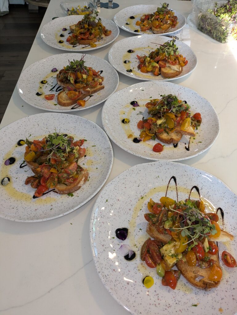 Eight plates of bruschetta topped with colorful cherry tomatoes, microgreens, and drizzles of balsamic glaze are arranged on a white countertop.
