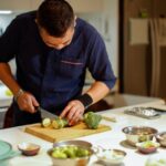 A man in a navy shirt focuses on slicing zucchini on a wooden cutting board in a modern kitchen. Bowls of vegetables surround him, creating a culinary atmosphere.