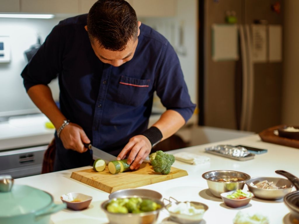 A man in a navy shirt focuses on slicing zucchini on a wooden cutting board in a modern kitchen. Bowls of vegetables surround him, creating a culinary atmosphere.